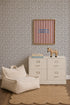 A modern nursery featuring Sprinkle Wallpaper in black, a white dresser, a plush chair, a scalloped beige rug, a toy zebra, books, and striped wall art with blue text.