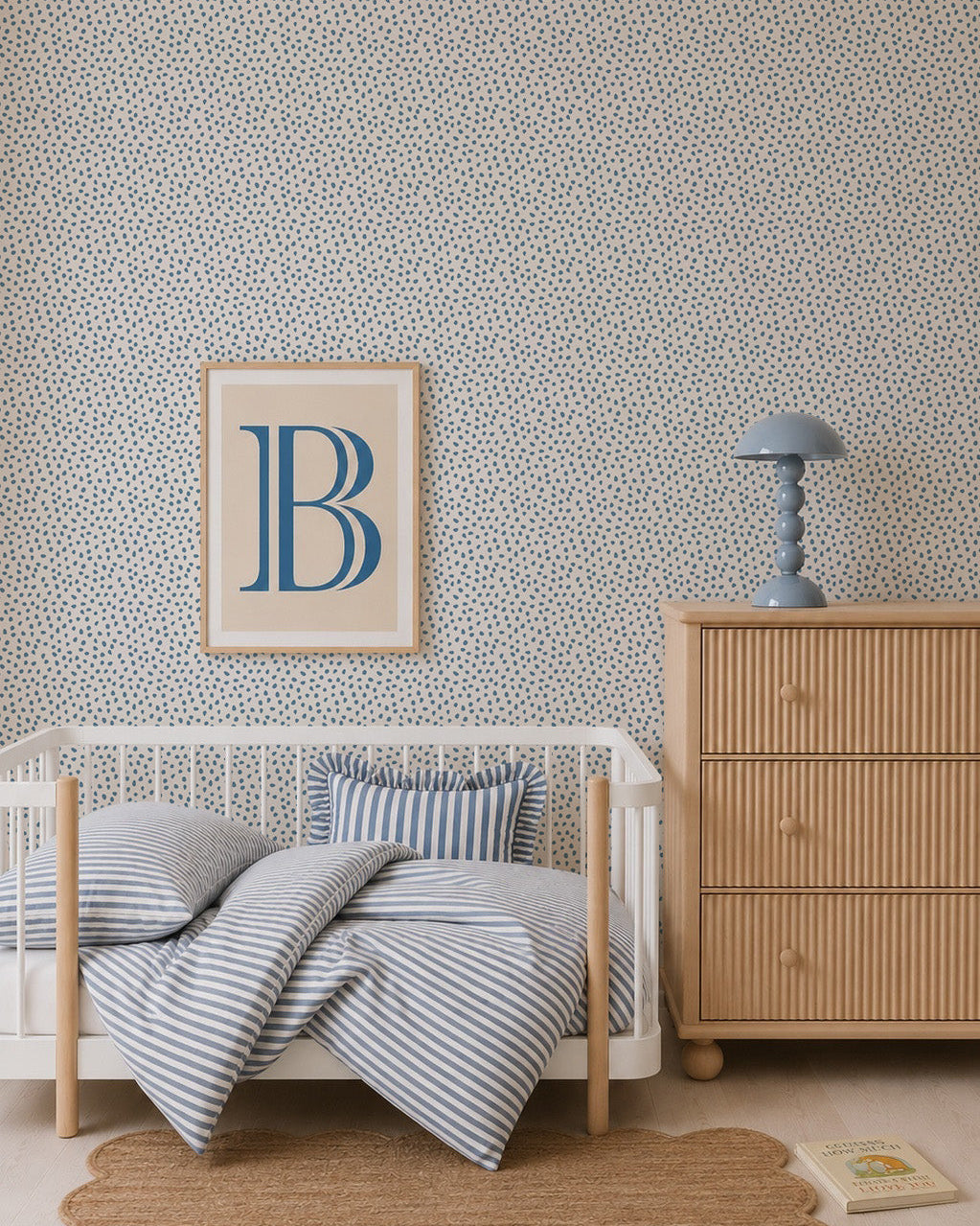A cozy nursery features a white crib, blue striped bedding, wooden dresser, blue lamp, framed art with a large blue B, a round woven rug, and Sprinkle Wallpaper in Blue. A book lies on the floor to complete the inviting scene.