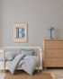 A cozy nursery features a white crib, blue striped bedding, wooden dresser, blue lamp, framed art with a large blue B, a round woven rug, and Sprinkle Wallpaper in Blue. A book lies on the floor to complete the inviting scene.