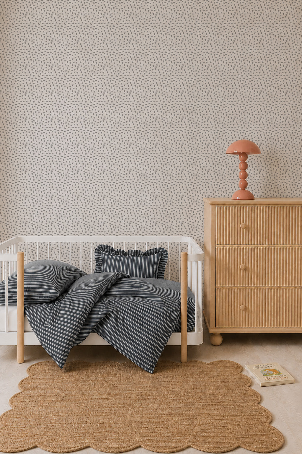 A minimal nursery with a white crib, striped bedding, a wooden dresser topped with a pink lamp, a scalloped rug, Sprinkle Wallpaper in Grey featuring a repeating pattern, and a book on the floor showcases thoughtful interior design.