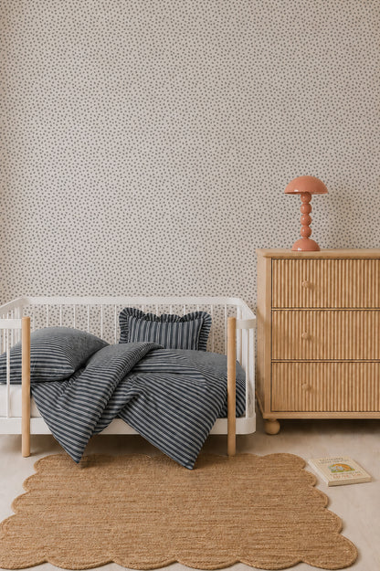 A minimal nursery with a white crib, striped bedding, a wooden dresser topped with a pink lamp, a scalloped rug, Sprinkle Wallpaper in Grey featuring a repeating pattern, and a book on the floor showcases thoughtful interior design.