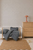 A minimal nursery with a white crib, striped bedding, a wooden dresser topped with a pink lamp, a scalloped rug, Sprinkle Wallpaper in Grey featuring a repeating pattern, and a book on the floor showcases thoughtful interior design.