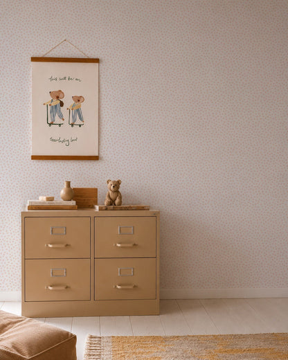 A beige dresser with four drawers stands against a wall decorated in Sprinkle Wallpaper Sample, Pink. On top, a teddy bear, stacked books, and small decor are displayed. Above hangs a framed print featuring two figures and handwritten text.