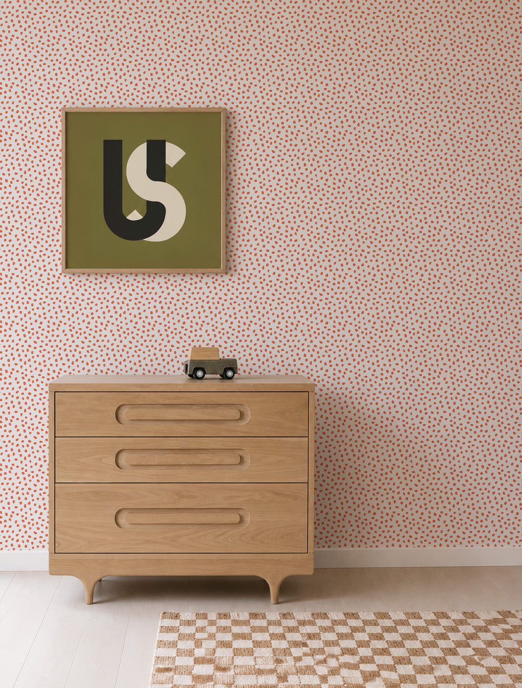 A light wood dresser with three drawers stands against a wall adorned in Sprinkle Wallpaper, Cherry Red, featuring playful pink dotted sprinkles. Above it hangs abstract art in a green frame. A small toy car sits atop the dresser, while a beige checkered rug rests on the pale floor.