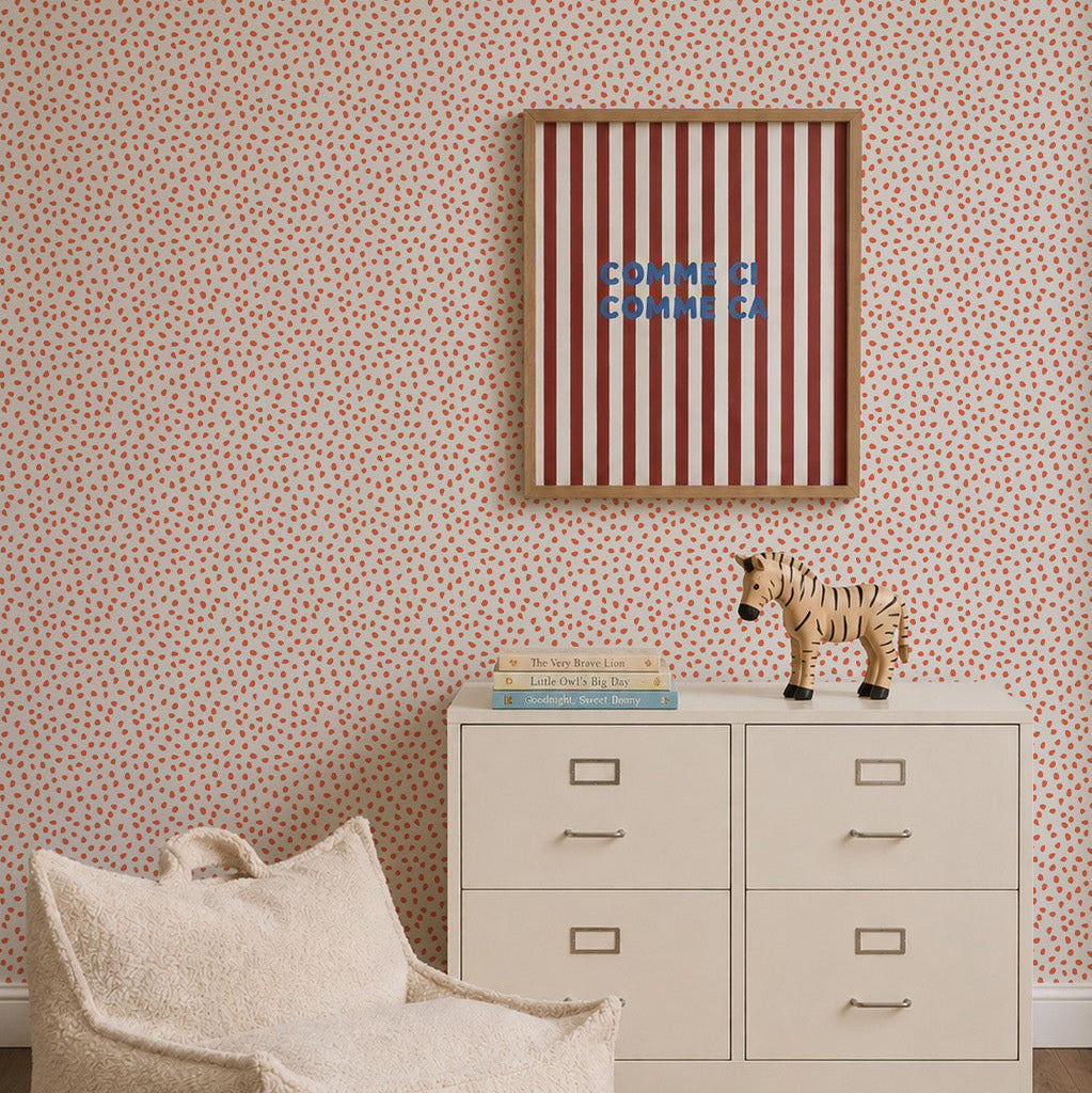 A small dresser with books and a zebra figurine stands against a statement wall featuring the Sprinkle Wallpaper in Cherry Red. Above the dresser, a striped artwork with blue text is displayed. A cream beanbag chair sits in the foreground.