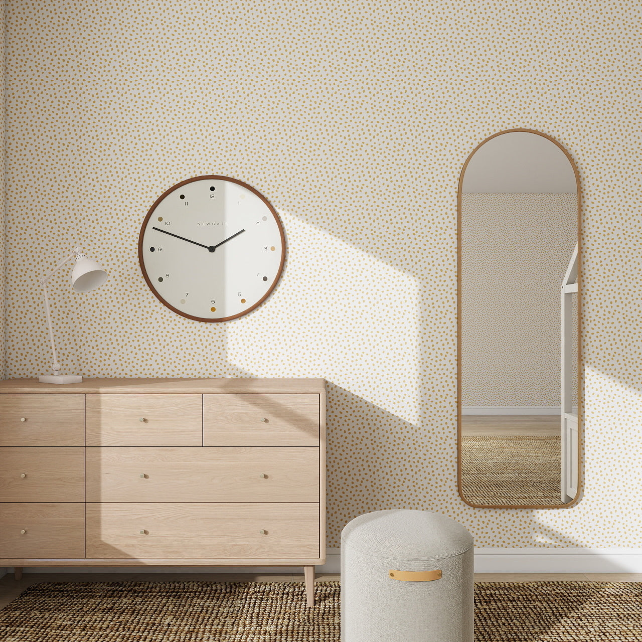A minimalist room features a statement wall with Sprinkle Wallpaper Repeat Pattern - Mustard, a round clock, oval mirror, wooden dresser, white desk lamp, padded stool, and woven flooring. Sunlight casts shadows on the wall and floor.