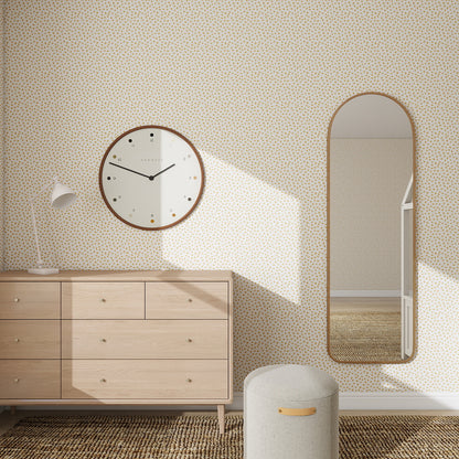 A minimalist room features a statement wall with Sprinkle Wallpaper Repeat Pattern - Mustard, a round clock, oval mirror, wooden dresser, white desk lamp, padded stool, and woven flooring. Sunlight casts shadows on the wall and floor.