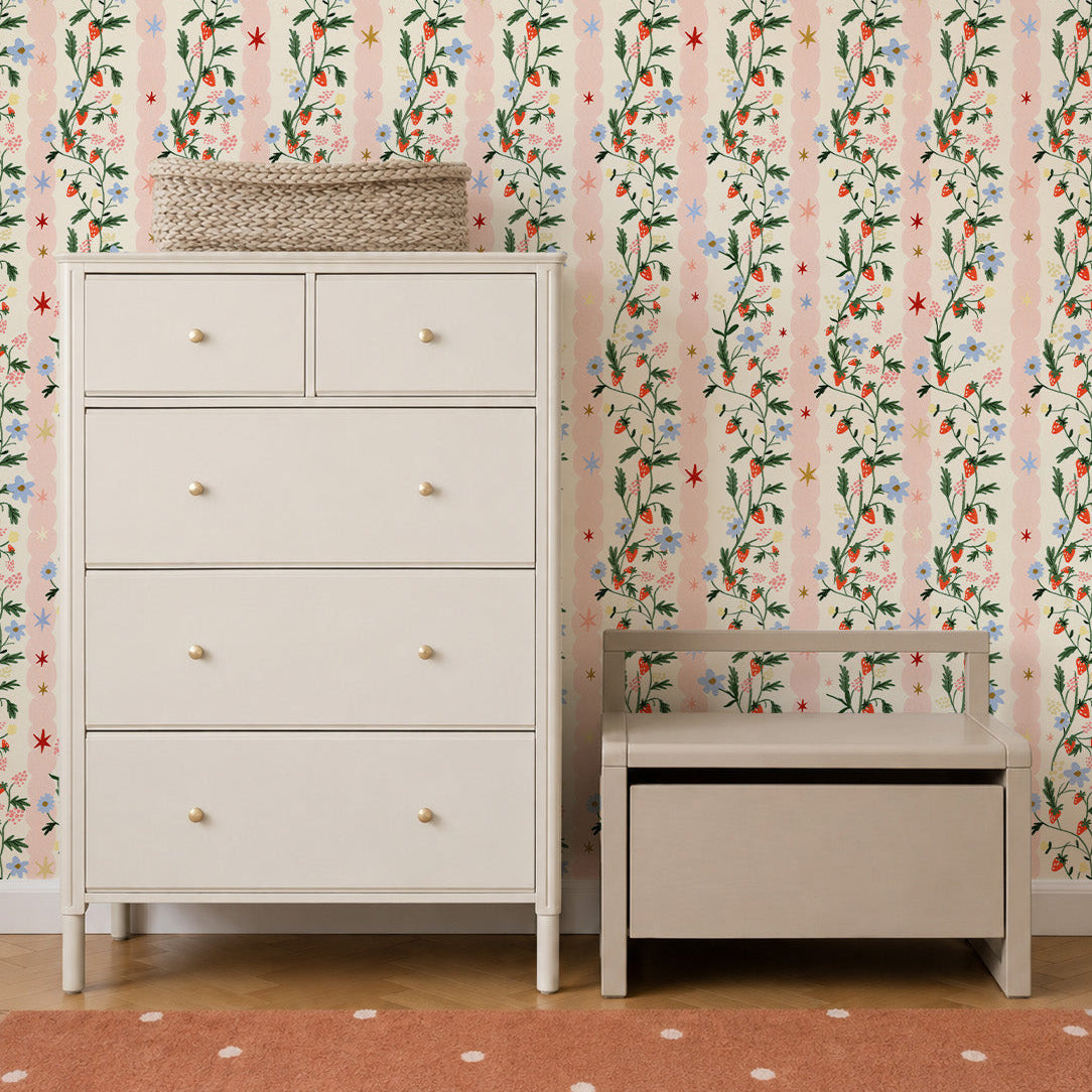 A white dresser and small wooden storage bench sit against a wall featuring Strawberry Bloom Wallpaper Sample with vertical pink stripes, strawberries, blue flowers, and stars.