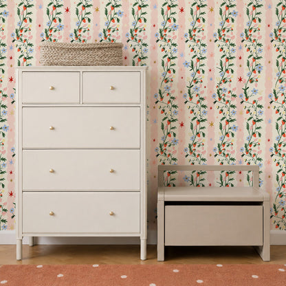 A white dresser and small wooden storage bench sit against a wall featuring Strawberry Bloom Wallpaper Sample with vertical pink stripes, strawberries, blue flowers, and stars.
