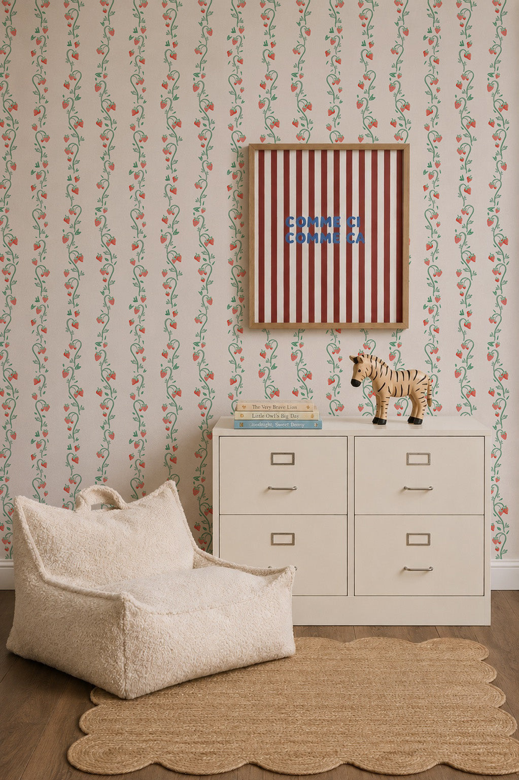 A cozy kids room featuring Strawberry Stripes Wallpaper, a white dresser topped with books and a tiger figurine, a soft cream chair, and a scalloped beige rug. A framed, striped Esther sign hangs above the dresser.