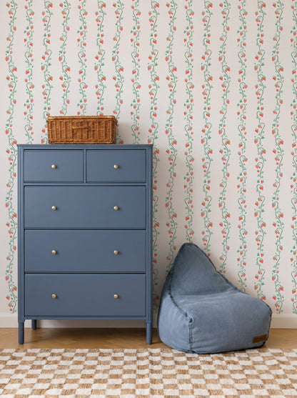 A blue dresser and blue bean bag sit on a checkered rug in front of charming Strawberry Stripes Wallpaper. A wicker basket is on top of the dresser, creating a cozy and neatly arranged scene.