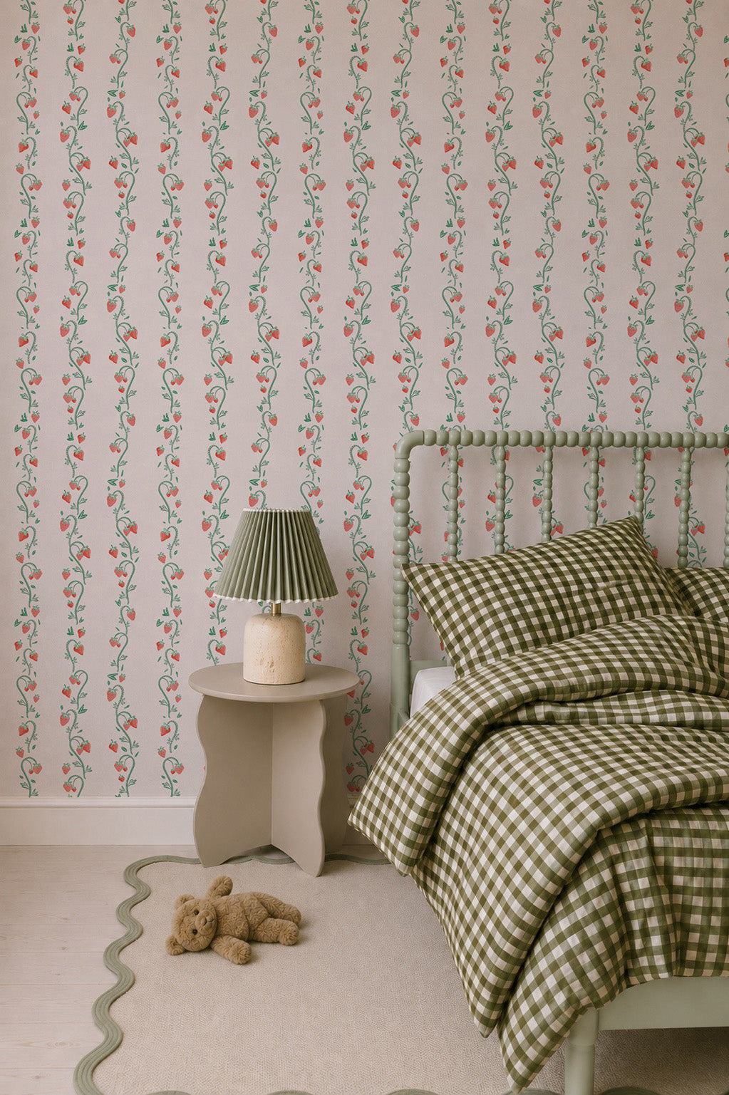Cozy bedroom with green gingham bedding on a spindle bed, Strawberry Stripes Wallpaper in the kids’ room, a wavy-edged nightstand with a green lamp, beige rug with a scalloped edge, and a teddy bear on the floor.