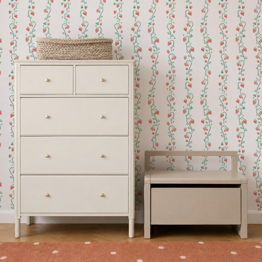 A cream-colored dresser and matching bench sit on a wooden floor with a peach polka dot rug. The wall behind features Strawberry Stripes Wallpaper. A woven basket is on top of the dresser.