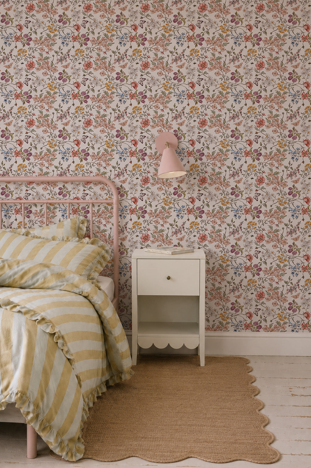A cozy bedroom features Sunlit Butterfly Meadow Wallpaper, a pink metal bed with yellow and white striped bedding, a small white nightstand, a pink wall lamp, and a tan woven rug on a white wooden floor.