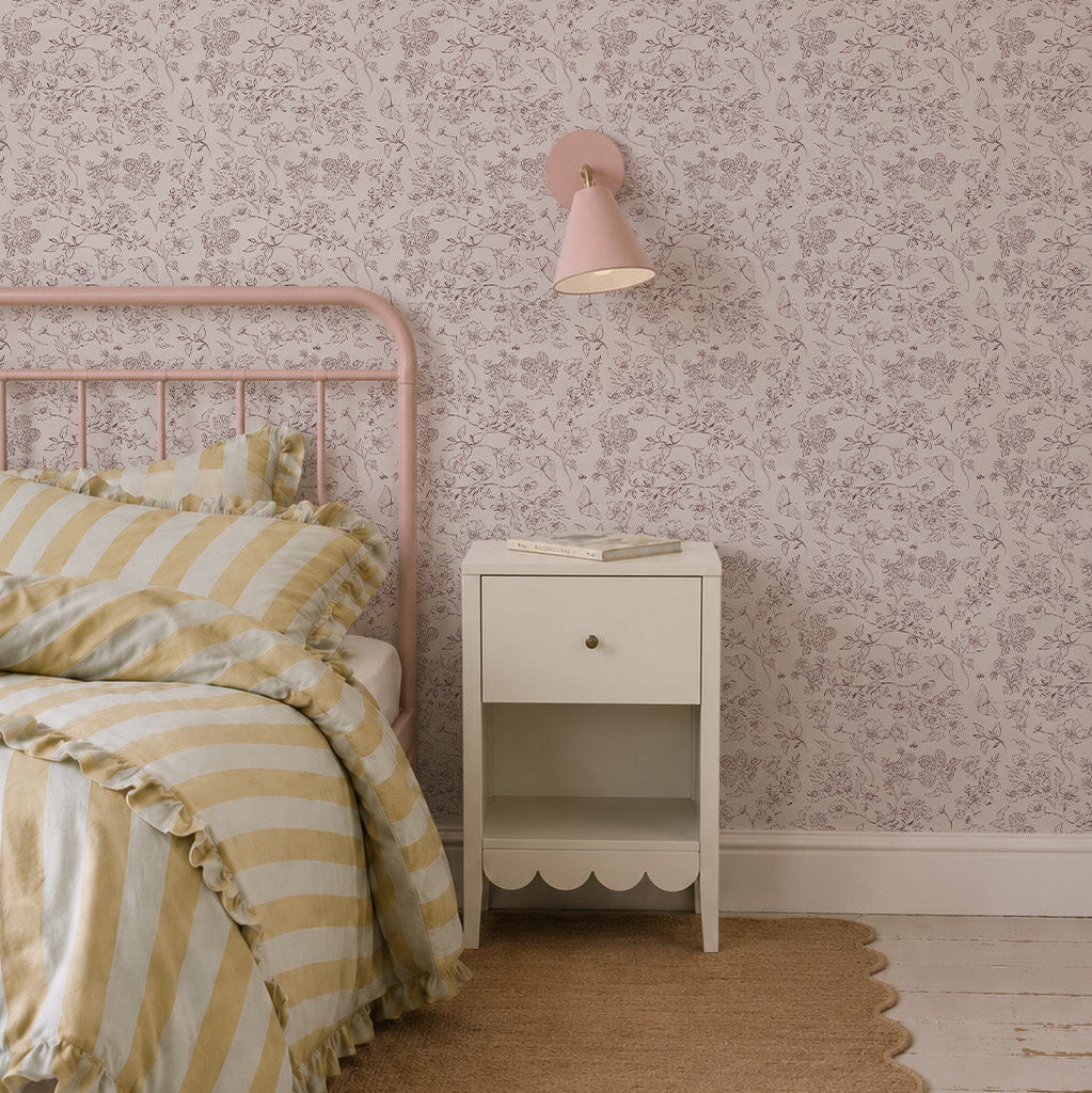 A pastel bedroom featuring a pink metal bed, striped yellow and white bedding, a small white nightstand with a book, and Sunlit Meadow Wallpaper with a pink wall sconce above the nightstand.