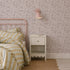 A pastel bedroom featuring a pink metal bed, striped yellow and white bedding, a small white nightstand with a book, and Sunlit Meadow Wallpaper with a pink wall sconce above the nightstand.