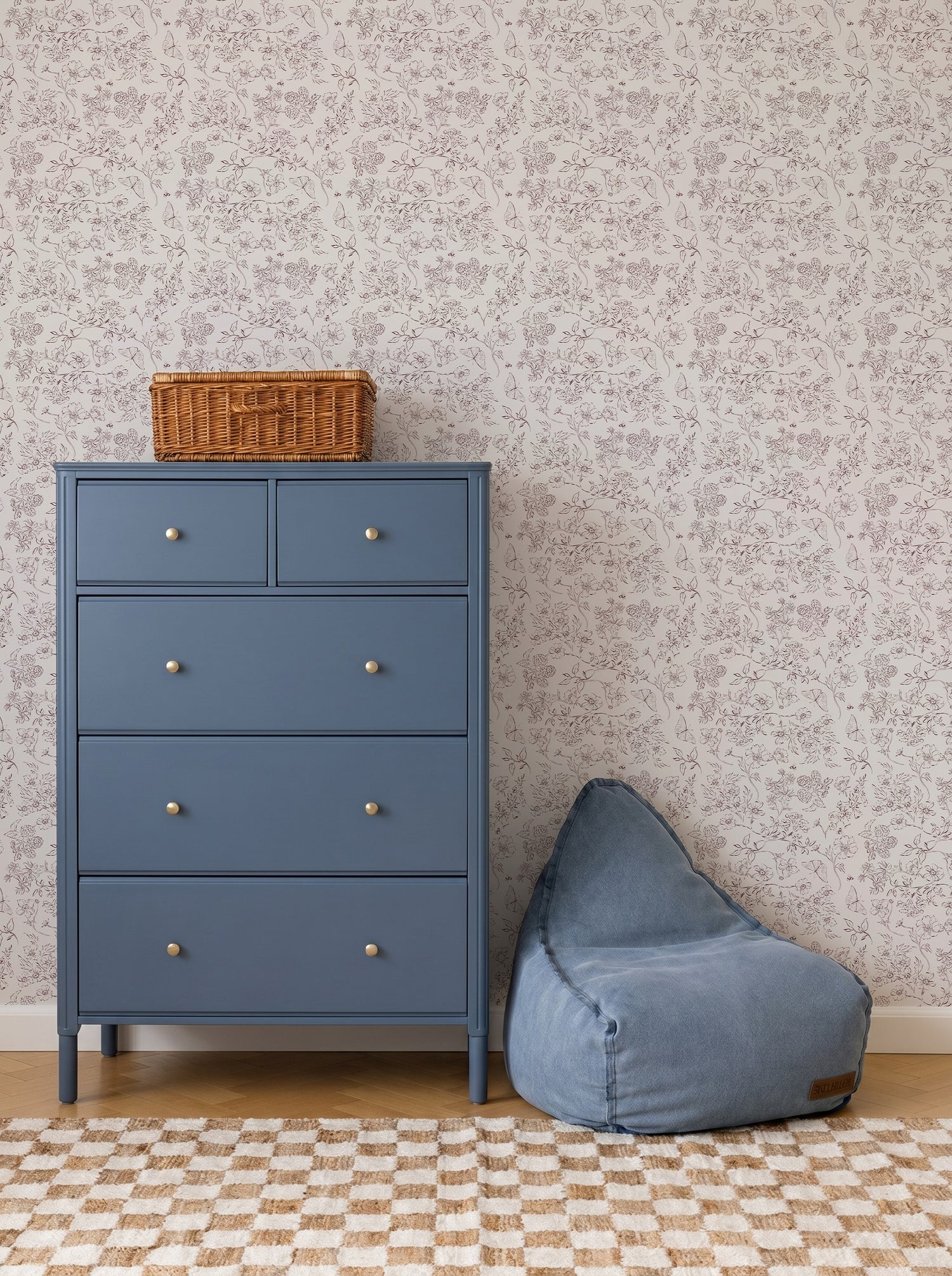 A blue dresser with five drawers stands next to a blue bean bag, topped with a wicker basket. The background features Sunlit Meadow Wallpaper and a checkered rug on a wooden floor.