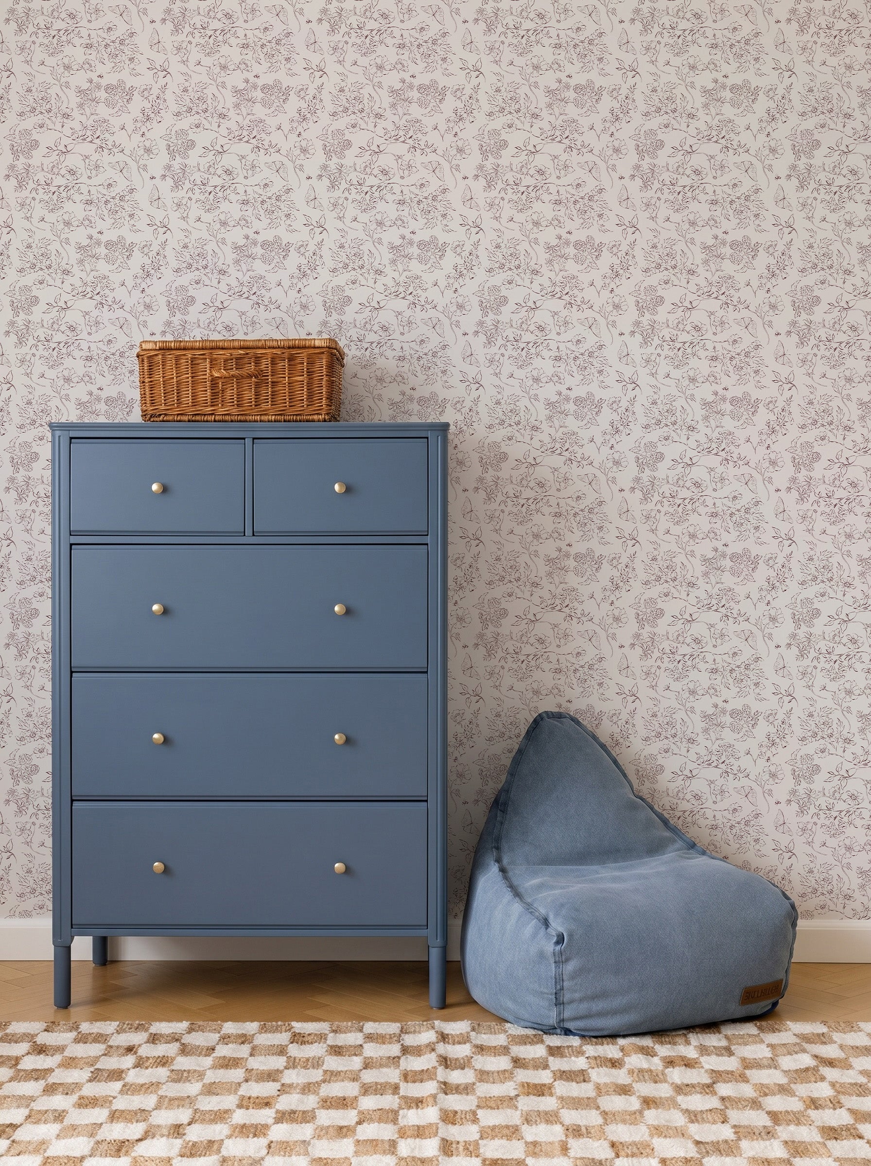A blue dresser with five drawers stands next to a blue bean bag, topped with a wicker basket. The background features Sunlit Meadow Wallpaper and a checkered rug on a wooden floor.