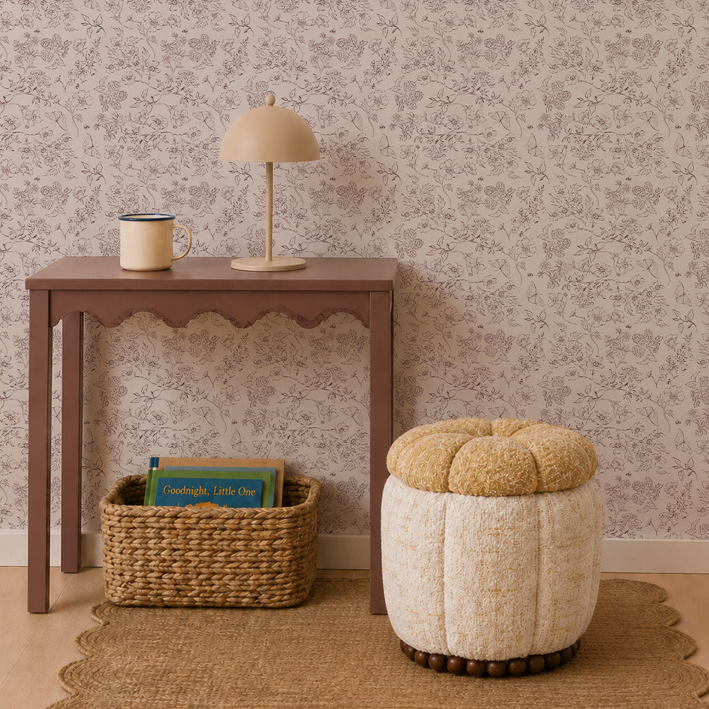 A small brown table with a lamp and mug, a woven basket filled with children’s books, and a round textured stool are arranged on a rug against Sunlit Meadow Wallpaper.
