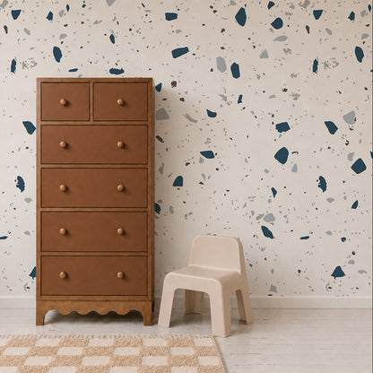 A tall brown dresser with six drawers stands next to a small beige chair on a white floor. Behind them, the wall is adorned with the Terrazzo Mural, Blue, featuring modern blue and gray shapes. A checkered beige rug is partially visible—making this space a stylish statement in home decor.