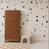 A tall brown dresser with six drawers stands next to a small beige chair on a white floor. Behind them, the wall is adorned with the Terrazzo Mural, Blue, featuring modern blue and gray shapes. A checkered beige rug is partially visible—making this space a stylish statement in home decor.