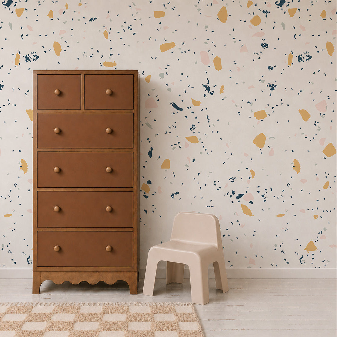 A tall brown wooden dresser and a small beige plastic chair sit on a light floor in front of a white wall adorned with the Terrazzo Mural Sample in Yellow. A beige and white checkered rug is partially visible.