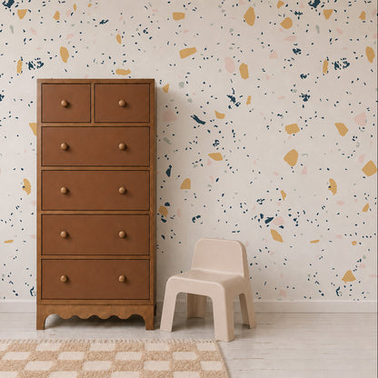 A tall brown wooden dresser and a small beige plastic chair sit on a light floor in front of a white wall adorned with the Terrazzo Mural Sample in Yellow. A beige and white checkered rug is partially visible.