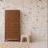 A tall brown wooden dresser and a small beige plastic chair sit on a light floor in front of a white wall adorned with the Terrazzo Mural Sample in Yellow. A beige and white checkered rug is partially visible.