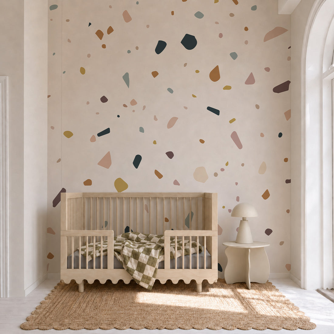 A neutral nursery features a wooden crib, a checkered blanket, and a small side table with lamp, with the Terrazzo Spritz Mural used as an accent wall. Natural light streams in from a tall arched window, highlighting the cozy and inviting atmosphere.