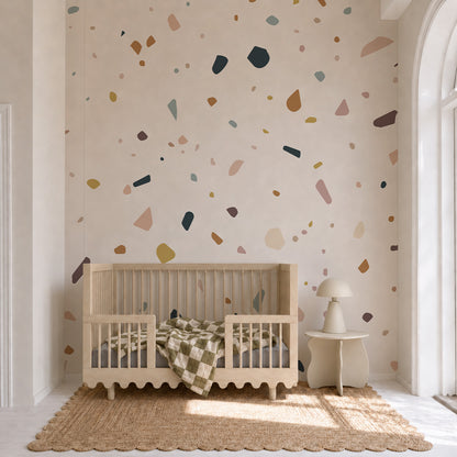 A neutral nursery features a wooden crib, a checkered blanket, and a small side table with lamp, with the Terrazzo Spritz Mural used as an accent wall. Natural light streams in from a tall arched window, highlighting the cozy and inviting atmosphere.