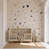 A neutral nursery features a wooden crib, a checkered blanket, and a small side table with lamp, with the Terrazzo Spritz Mural used as an accent wall. Natural light streams in from a tall arched window, highlighting the cozy and inviting atmosphere.