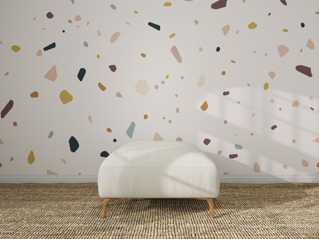 A white, rectangular ottoman rests on a woven rug before a wall decorated with Terrazzo Spritz Wallpaper Mural. Sunlight creates playful shadows, enhancing this minimalist setting—an ideal kids&