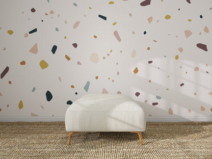 A white, rectangular ottoman rests on a woven rug before a wall decorated with Terrazzo Spritz Wallpaper Mural. Sunlight creates playful shadows, enhancing this minimalist setting—an ideal kids&