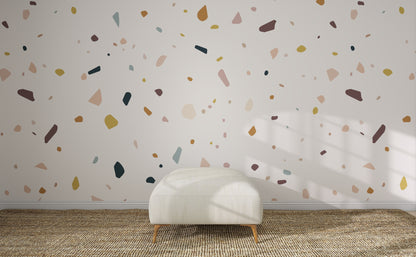 A white ottoman rests on a woven rug in front of a wall adorned with the Terrazzo Spritz Wallpaper Mural. Sunlight streams in, casting window shadows and bringing warmth to this lively, modern space.