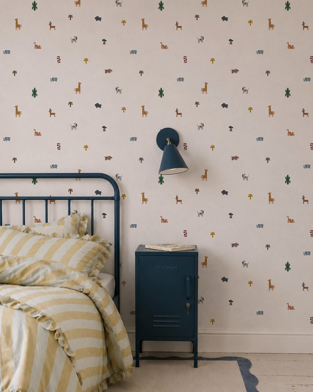 A cozy bedroom corner with a metal bed frame, yellow striped bedding, blue nightstand, wall-mounted lamp, and playful Tiny Tracks Safari Wallpaper—soft animal prints that add a whimsical touch to any kid’s room.