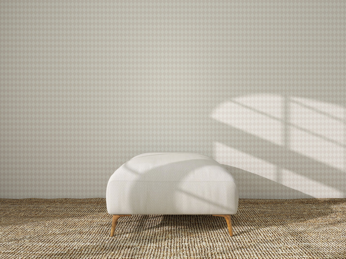 A white ottoman rests on a woven rug before a wall adorned with Diamond Dancer Wallpaper in Cream, as sunlight casts window-shaped shadows across the wall and floor.