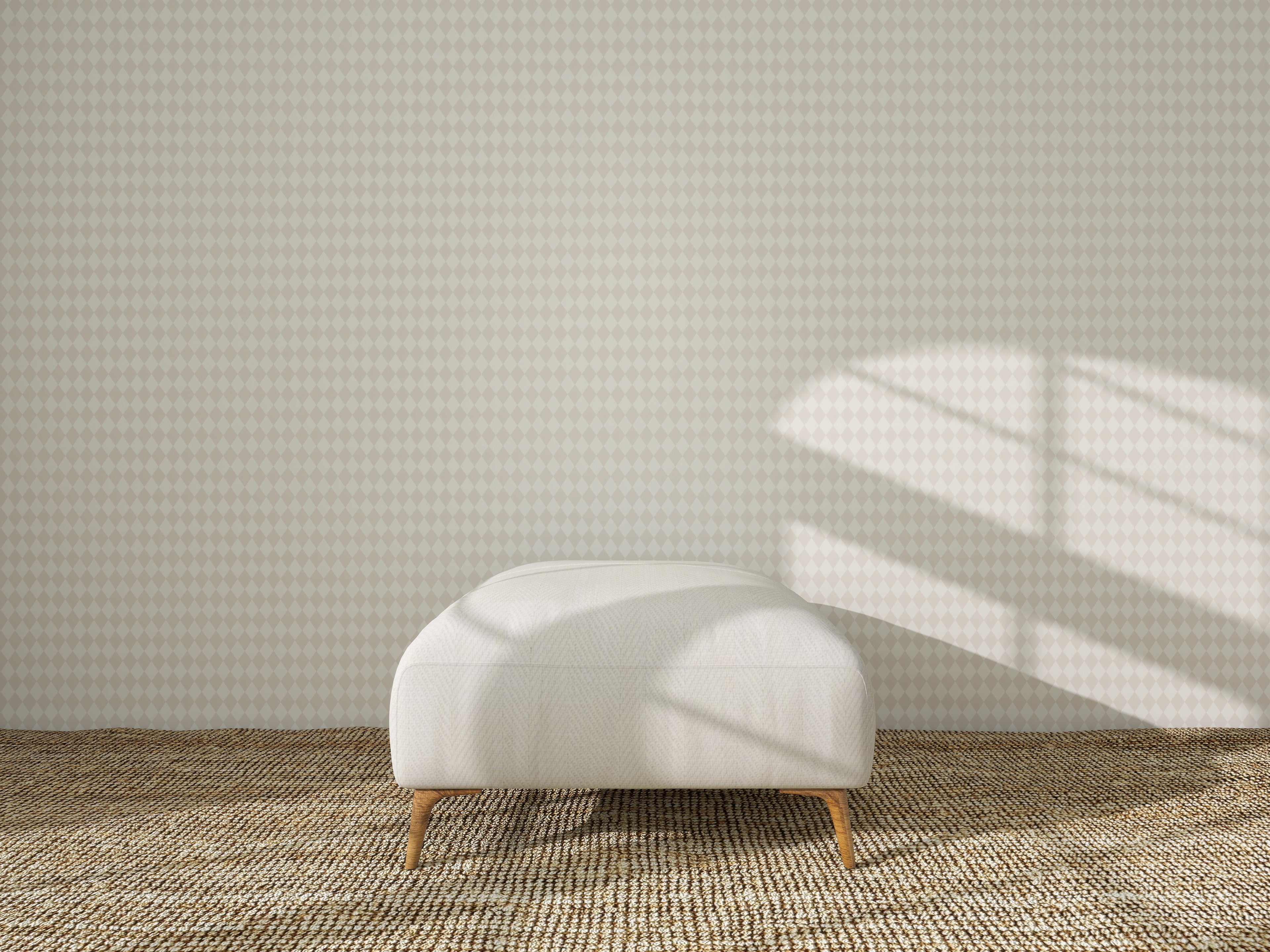A white ottoman rests on a woven rug before a wall adorned with Diamond Dancer Wallpaper in Cream, as sunlight casts window-shaped shadows across the wall and floor.