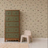 A green and wood dresser stands beside a small beige chair on a white floor with a checked rug. The wall features the Transport Trail Wallpaper in Beige, adorned with small colorful vehicles, creating a playful and inviting atmosphere in this charming kids’ room or nursery.