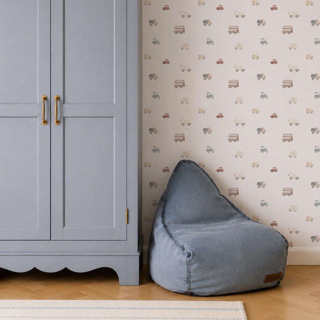 A soft blue bean bag sits by a light blue wardrobe with gold handles in this charming kids room. The wall behind is decorated with Transport Wallpaper featuring small cars, trucks, and bikes. A basket tops the wardrobe; part of a rug shows.