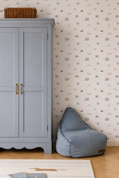 A soft blue bean bag sits by a light blue wardrobe with gold handles in this charming kids room. The wall behind is decorated with Transport Wallpaper featuring small cars, trucks, and bikes. A basket tops the wardrobe; part of a rug shows.