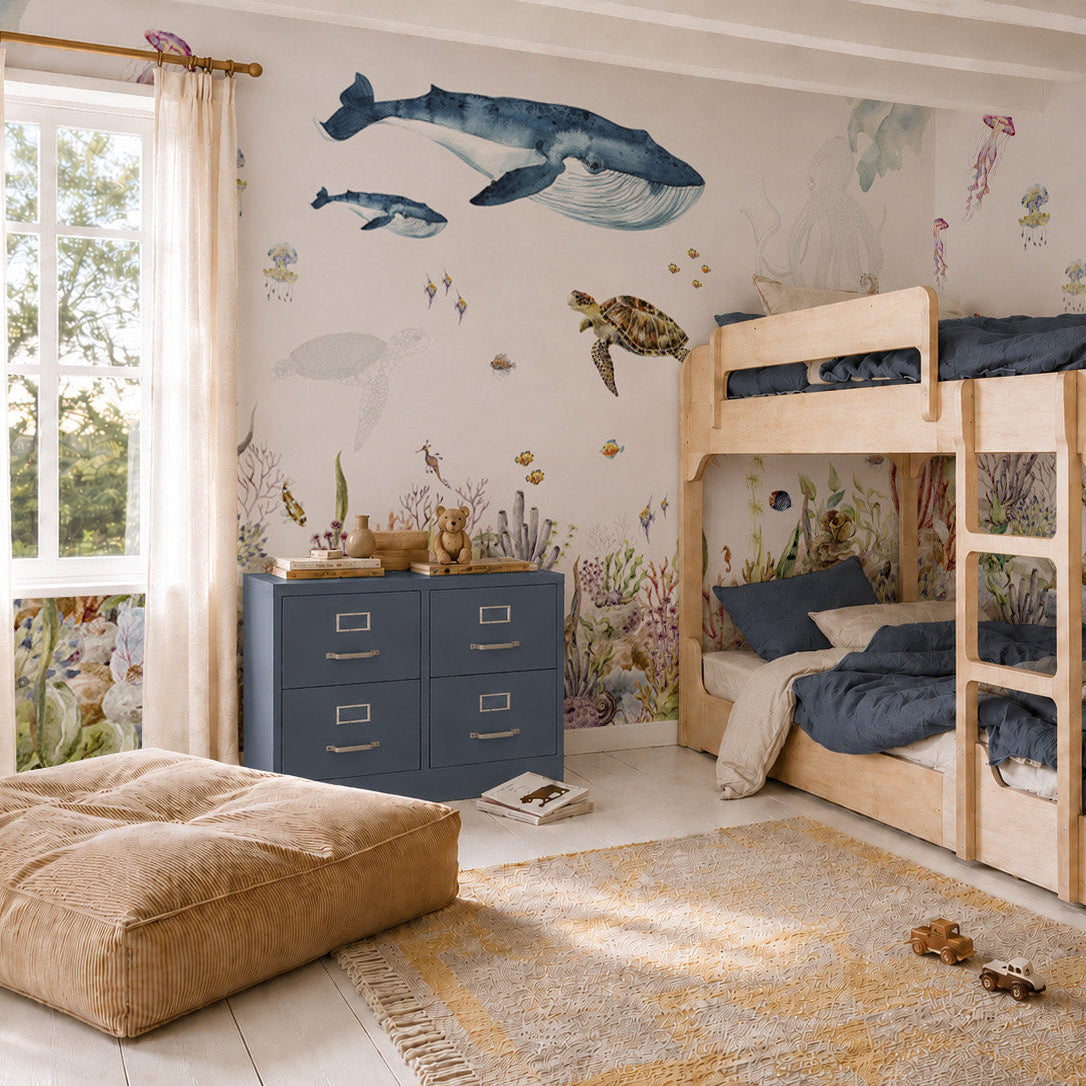 Children’s bedroom with wooden bunk beds, blue dresser, tan floor cushion, and an ocean mural featuring whales, fish, and a sea turtle above a vibrant underwater coral scene—perfect for ocean lovers. Sunlight streams through a window, highlighting the colorful Under The Sea Mural, Watercolour and playful sea creatures that bring this imaginative space to life.