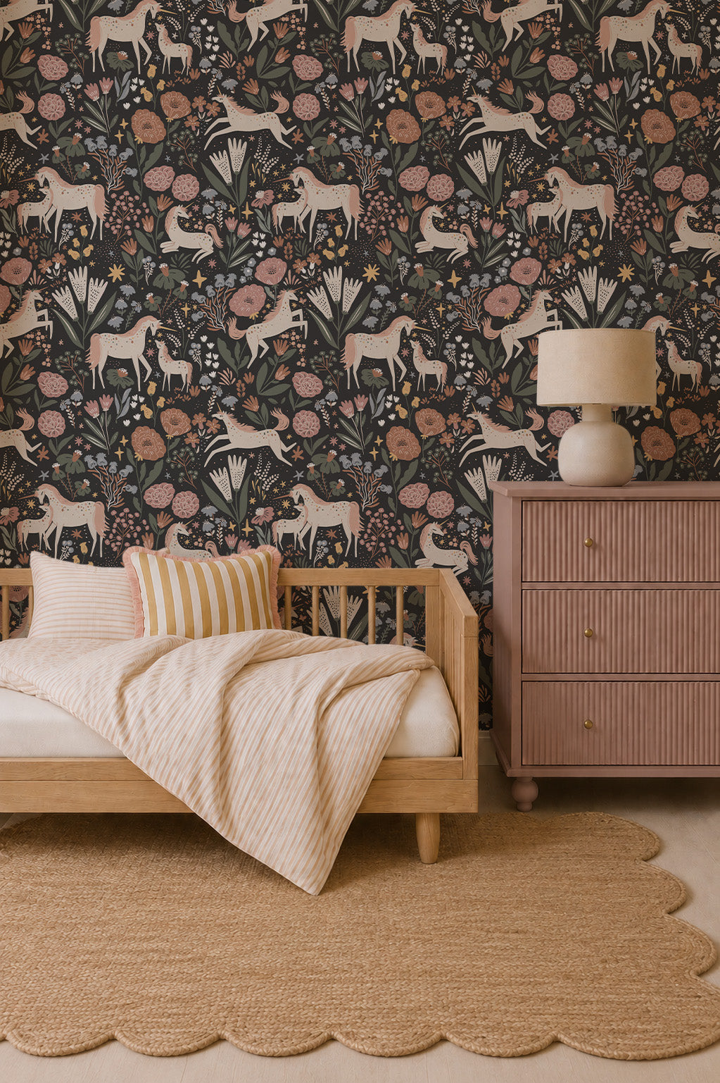A cozy bedroom with a wooden daybed, striped bedding, round rug, and a pink dresser topped with a lamp. The wall features whimsical design touches with Unicorn Meadow Wallpaper in Charcoal, featuring muted floral and horse patterns.