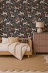 A cozy bedroom with a wooden daybed, striped bedding, round rug, and a pink dresser topped with a lamp. The wall features whimsical design touches with Unicorn Meadow Wallpaper in Charcoal, featuring muted floral and horse patterns.