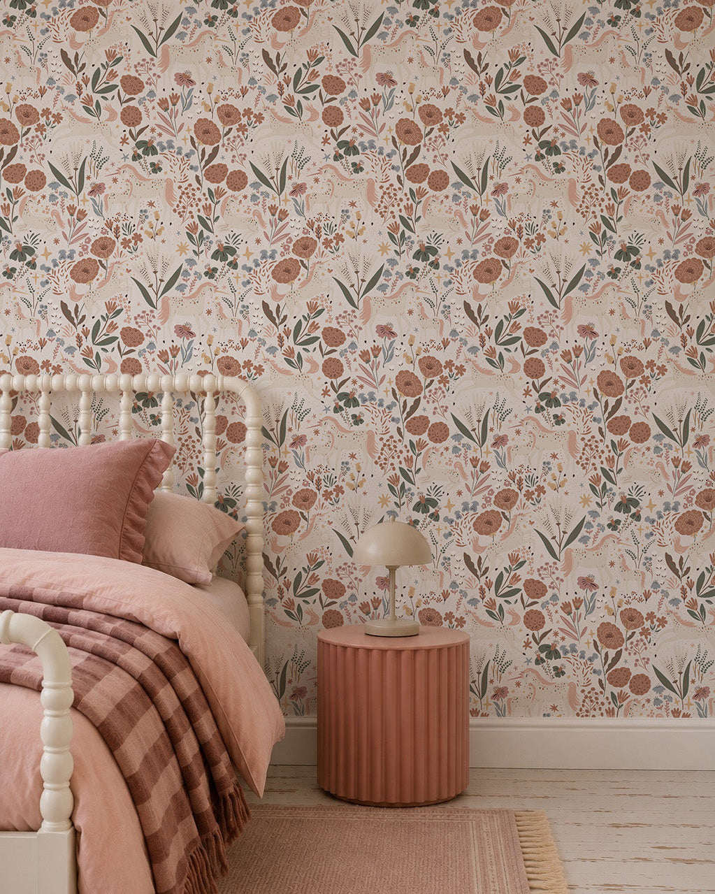 A cozy bedroom features a spindle bed with pink bedding, a plaid blanket, and a round ribbed nightstand topped with a beige lamp. The girly decor is enhanced by the Unicorn Meadow Wallpaper in cream.