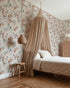 A cozy bedroom featuring Unicorn Meadow Wallpaper in Cream, a bed with a beige canopy and bedding, wooden stool, pendant light, woven rug, rattan cabinet, and wooden floor—perfect for girly room decor.