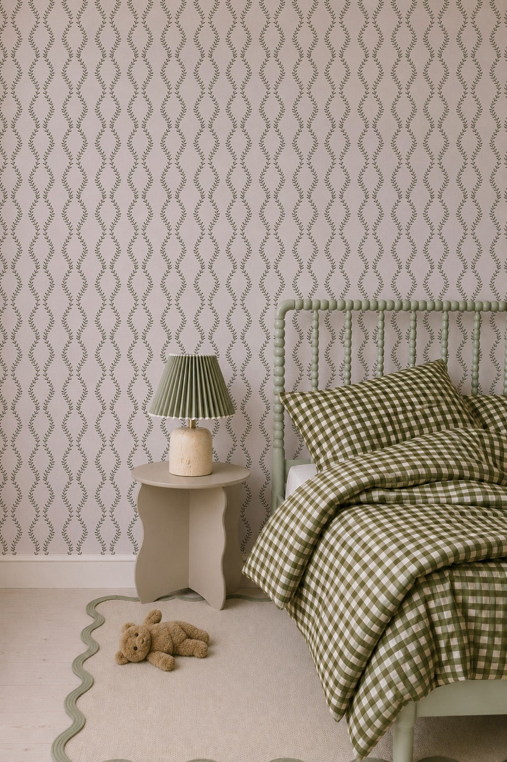 A cozy bedroom features a green checkered bedspread, a matching green bed frame, and a small table topped with a pleated green lamp for added charm. A beige rug with wavy edges and a teddy bear on the floor bring warmth to the space. Elegant Vintage Vines Wallpaper in soft tones provides an inviting backdrop, while hand-drawn details around the room enhance its nostalgic appeal against the patterned walls.