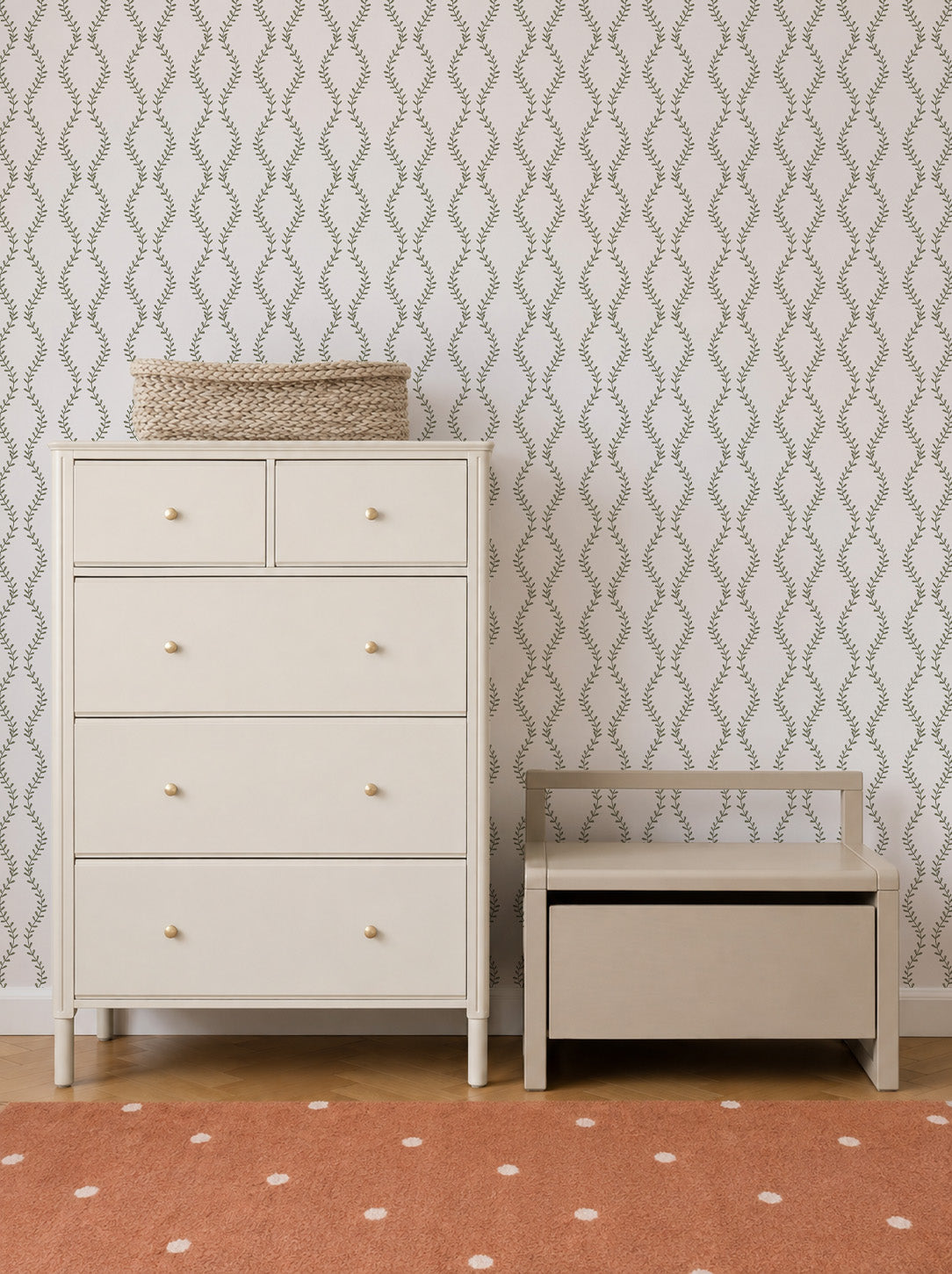 A cream-colored dresser with a woven basket on top stands beside a matching bench with storage. The wall features Vintage Vines Wallpaper, adding an elegant leafy pattern, and there’s an orange rug with white dots on the floor.