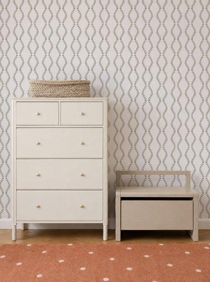 A cream-colored dresser with a woven basket on top stands beside a matching bench with storage. The wall features Vintage Vines Wallpaper, adding an elegant leafy pattern, and there’s an orange rug with white dots on the floor.