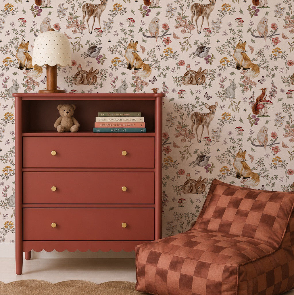 A children’s room with a terracotta dresser, books, a teddy bear, a lamp, a checker-patterned bean bag chair, a round jute rug, a plush bunny toy, and enchanting Whimsical Woodland Wallpaper that brings the magic of nature indoors.