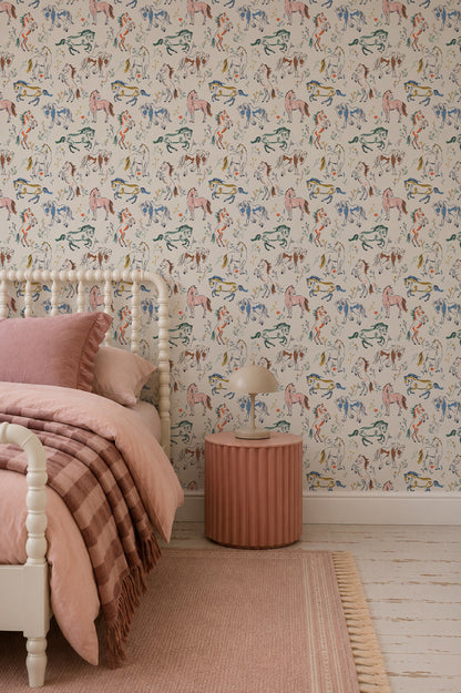 A bedroom featuring the Wild At Heart Wallpaper with vibrant, colorful hand-drawn horses sets a whimsical tone. The pink bed is dressed in a striped pink blanket and complemented by a round pink nightstand, beige lamp, and soft pink-toned rug and decor—creating a cozy, playful retreat ideal for children.